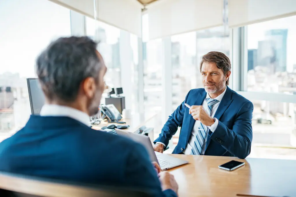 image of two men in suits sitting on opposite sides of a desk engaged in conversation