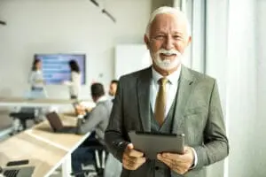 Senior male businessman holding digital tablet computer inside modern office with his colleagues.