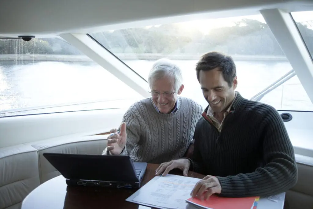 Father and son wearing sweaters and smiling as they look over documents together while sitting in a boat on the water