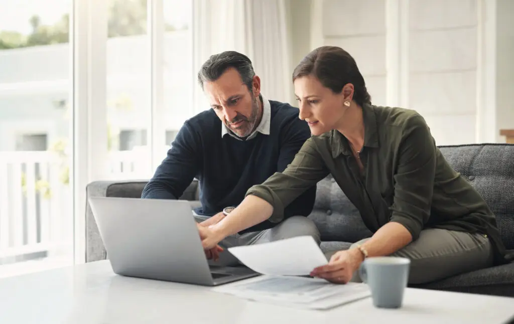 Couple sitting on a gray couch while looking at a laptop and documents on a coffee table