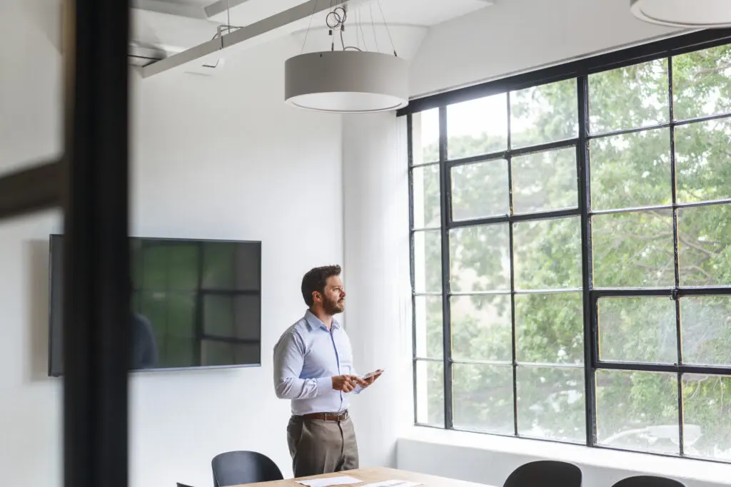 Business man in a light blue top and brown pants holding a tablet while looking out the window of a modern white office with large window