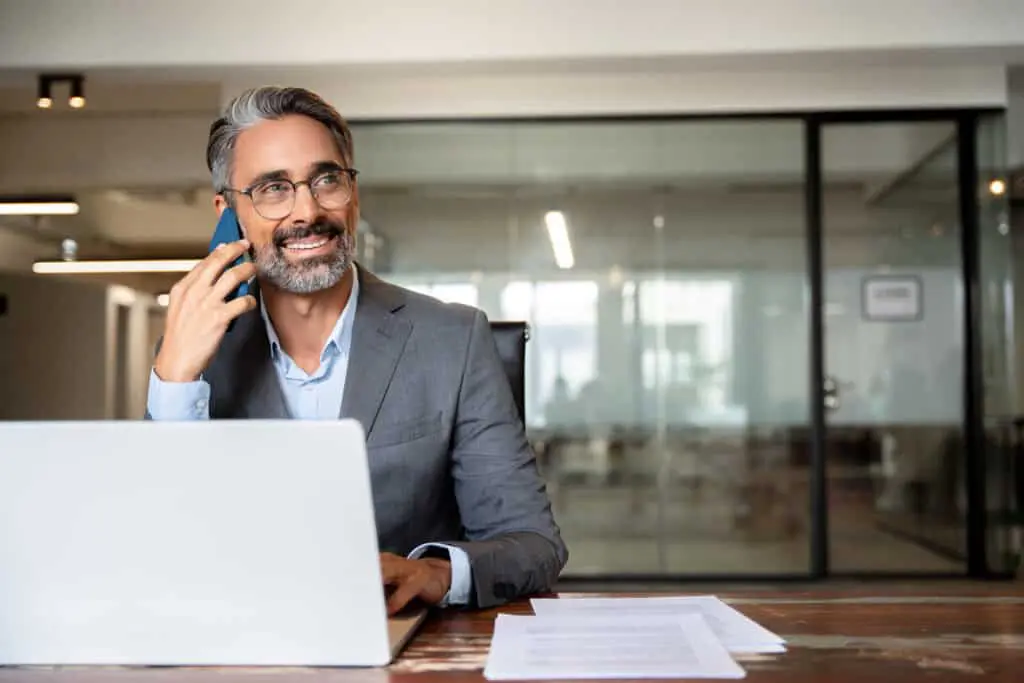 Middle aged Latin or Indian businessman having call on smartphone with business partners or clients. Smiling mature Hispanic man sitting at table talking by mobile cellphone at workplace in office.