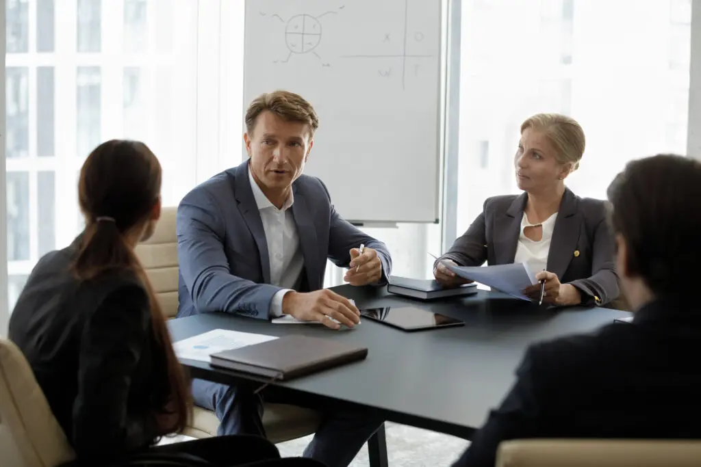 Middle-aged boss in suit lead corporate meeting with staff members sit at desk in modern office boardroom, search business solutions, take part in negotiations or group seminar event. Coaching concept