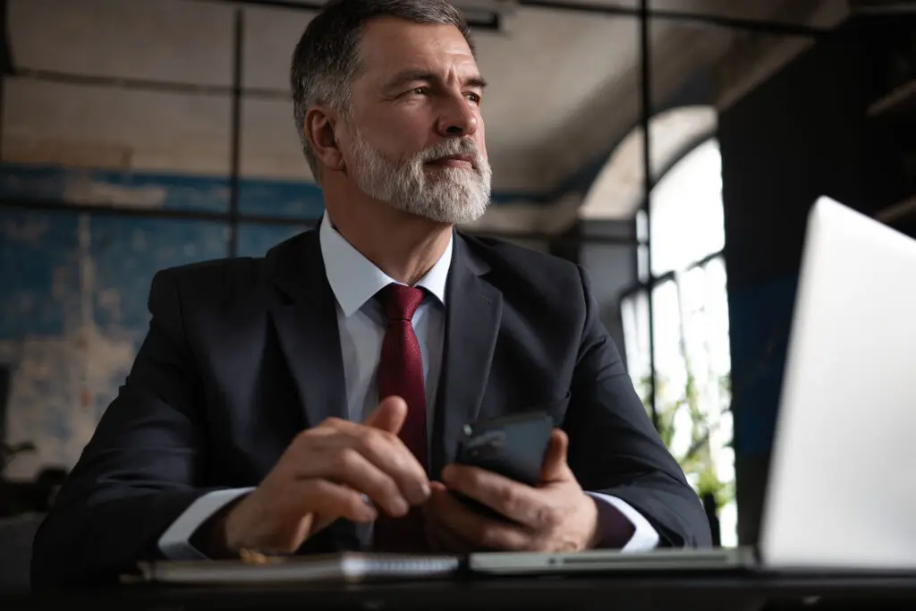 Mature business man in formal clothing using mobile phone. Serious businessman using smartphone and laptop at work