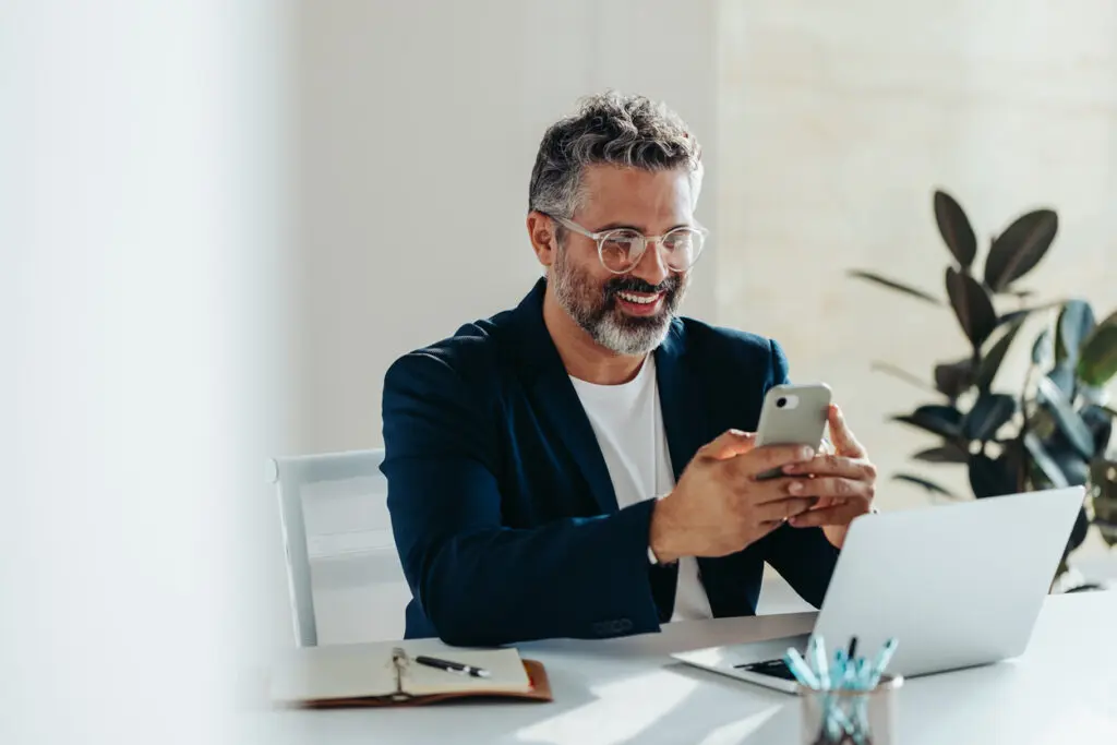 Cheerful, mature professional man is engrossed in his smartphone while sitting at a white desk in a sunny, contemporary office space. His laptop and notepad are at hand, suggesting productivity and connectivity.