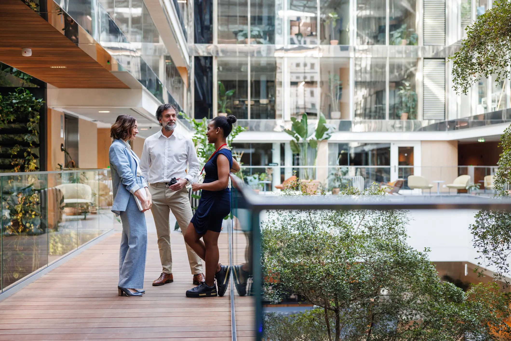 A group of diverse executives engages in a lively conversation in a sunlit office hallway, clad in professional attire, with digital tablets in hand.