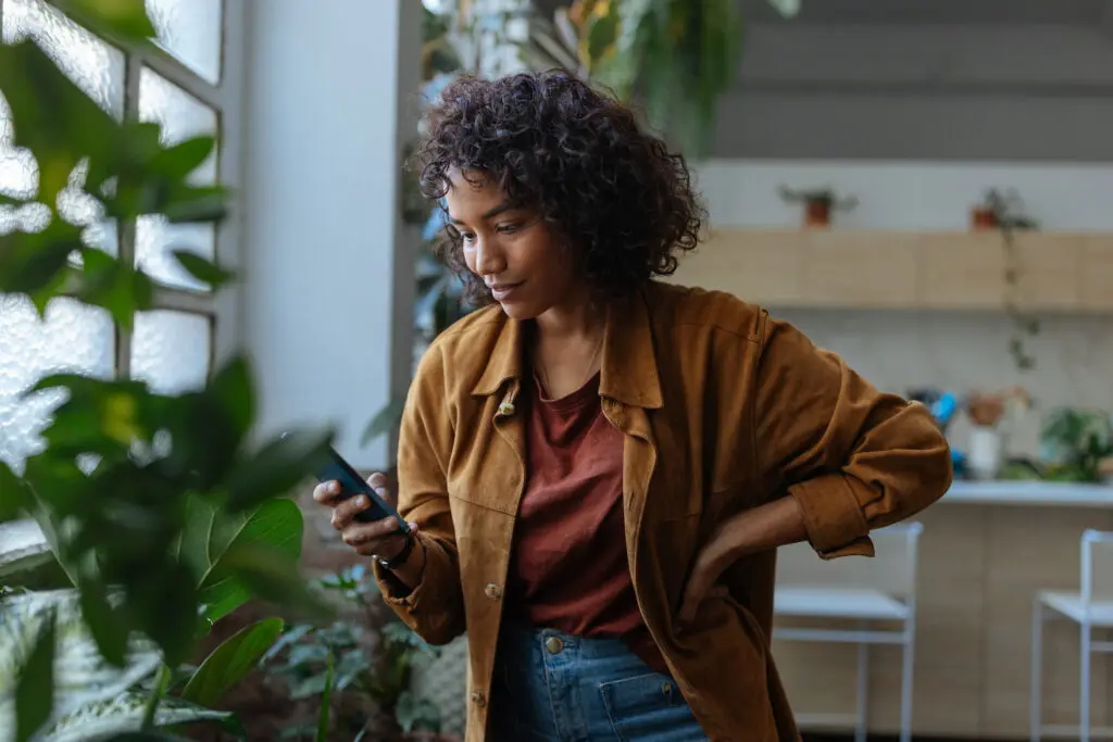 Young woman peacefully using her smartphone by a window in a cozy apartment, surrounded by green plants, radiating calm and happiness. Insured Cash Sweep page.