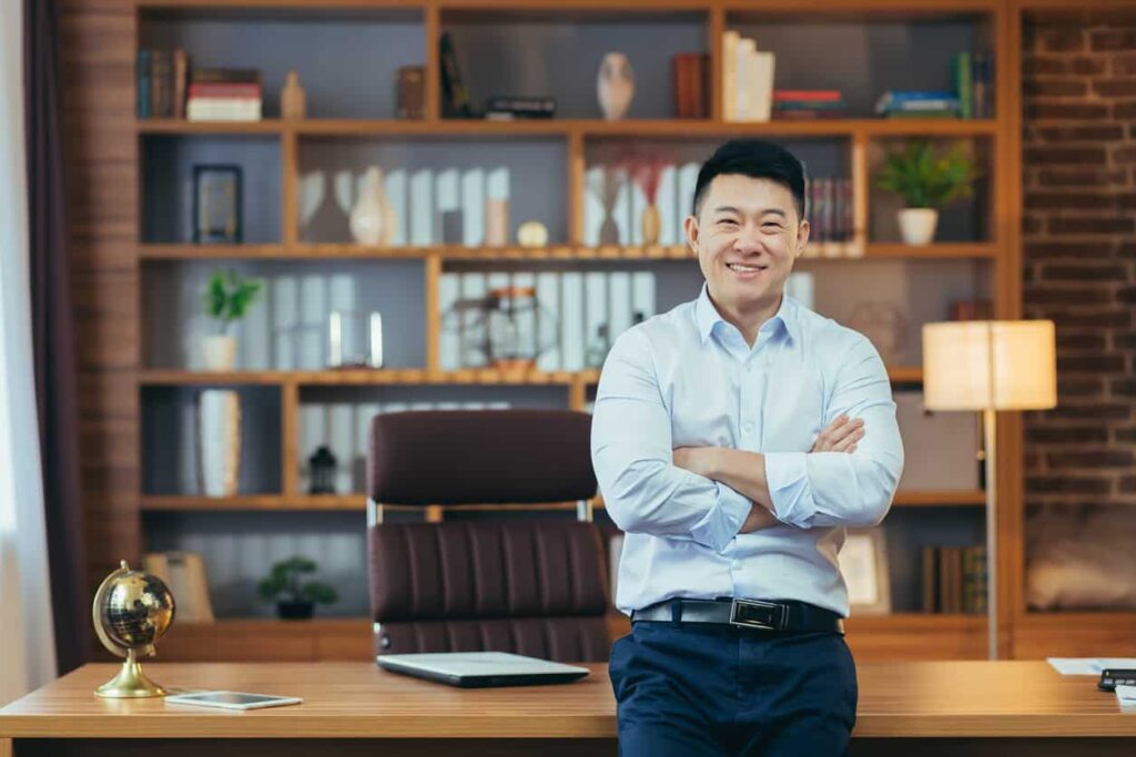 Asian business man smiling with arms crossed while leaning on a desk with his office behind him