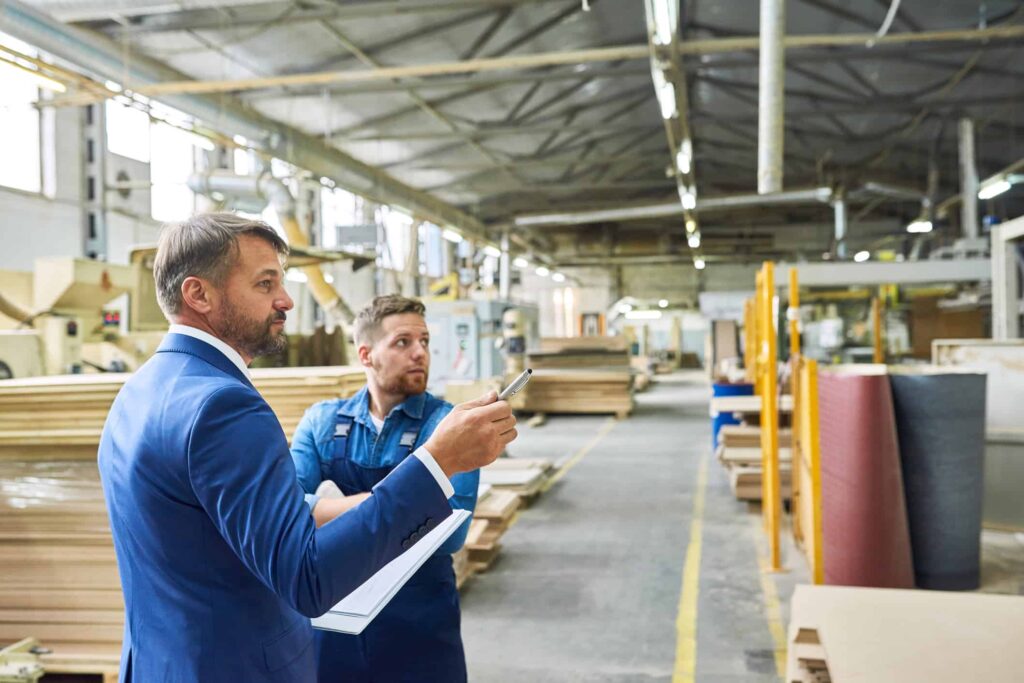 Business man in a blue suit holding a pen and pointing to something in a warehouse while talking with an employee in an apron