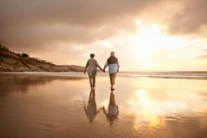 Elderly couple walking on the beach during low tide with the sunset and houses and rocks in the distance