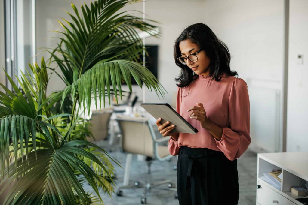 Business woman with glasses looking at an ipad in a modern office with a green plant to her right