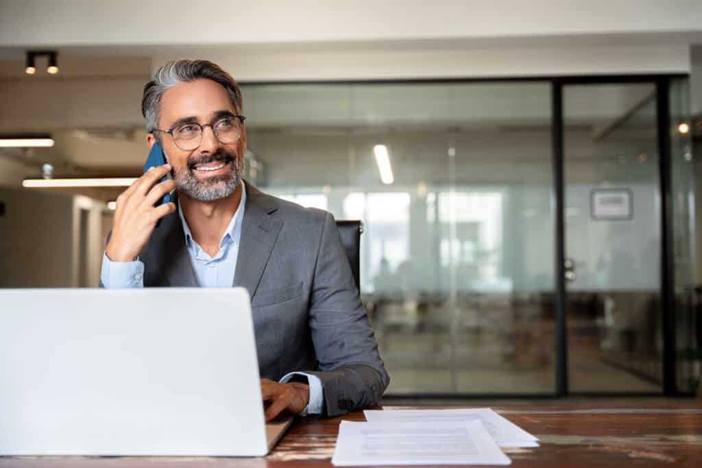 Middle aged Latin or Indian businessman having call on smartphone with business partners or clients. Smiling mature Hispanic man sitting at table talking by mobile cellphone at workplace in office.