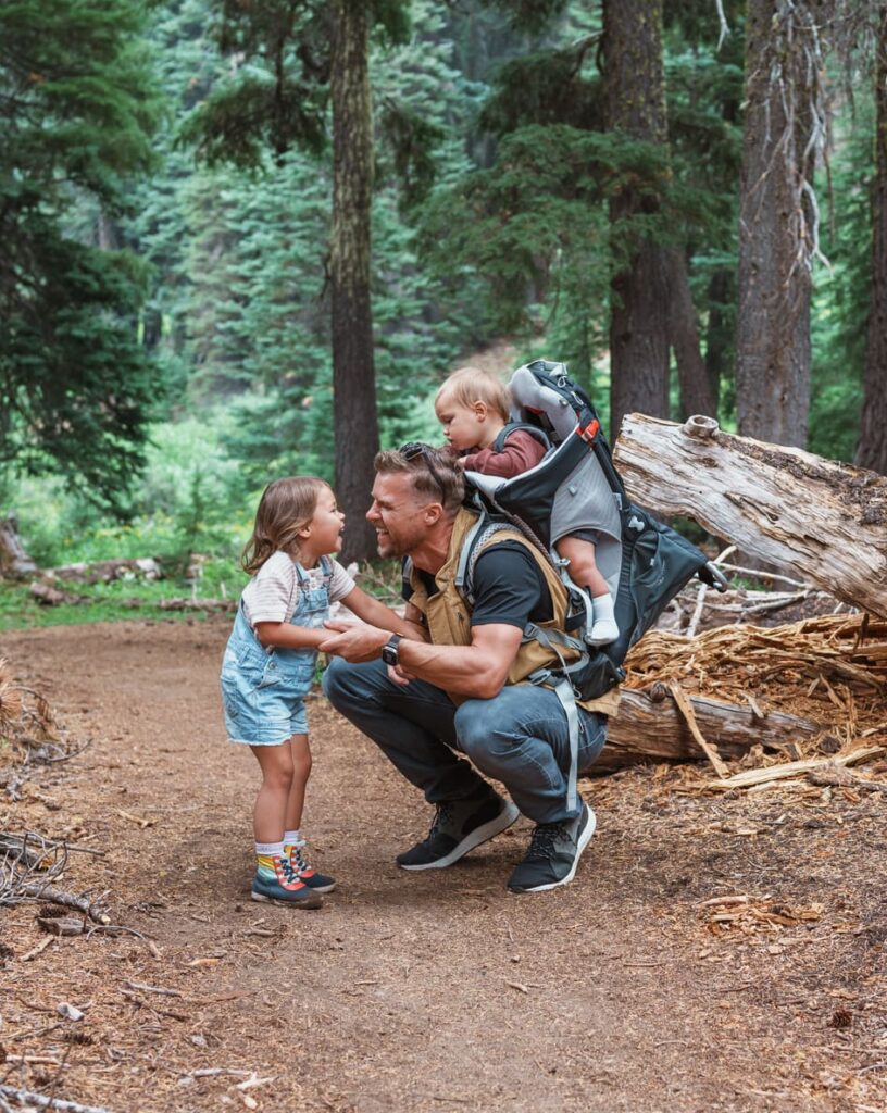 Young Caucasian father carrying his Eurasian one year old son while crouching down to talk to young daughter, who is excited to talk to him during a family hike in Oregon.