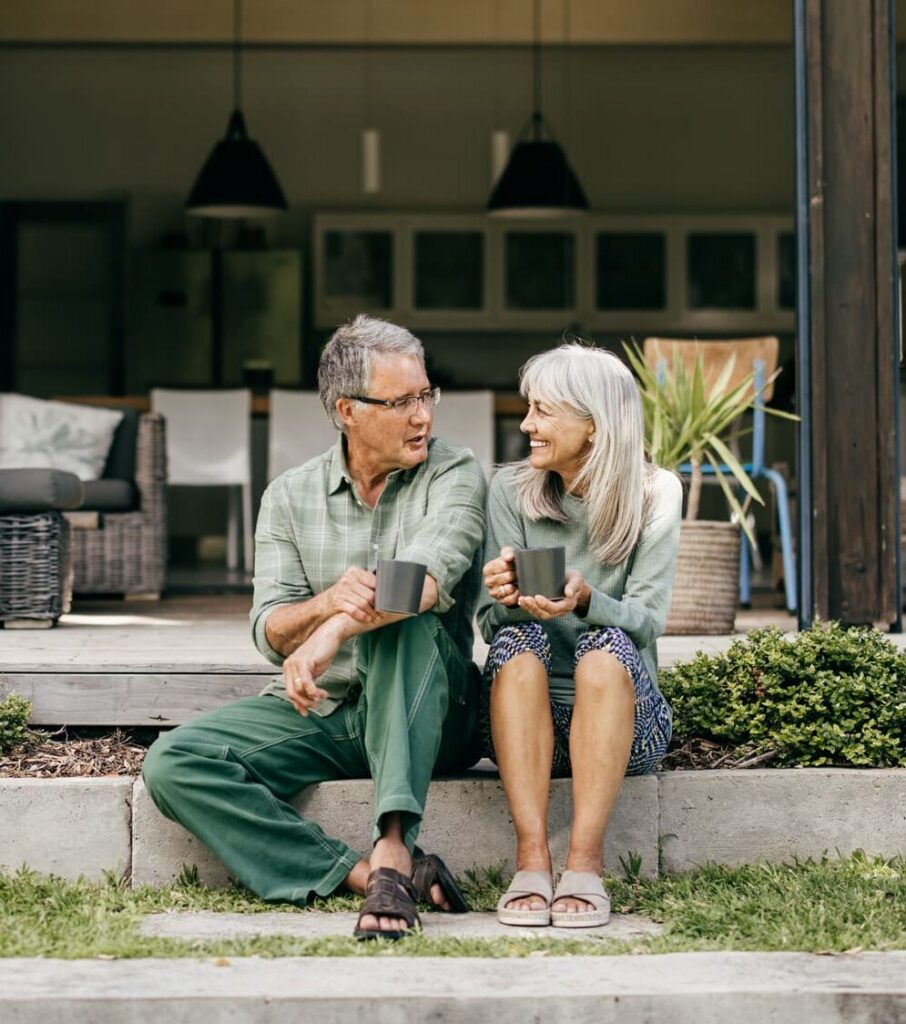 A joyful couple in their 60s leans close, sharing a heartfelt moment with coffee in hand on their cozy patio.
